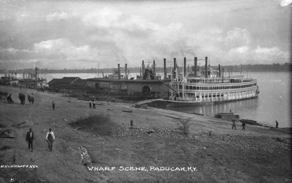 Elevated view of several steamboats docked at the wharf. Some of the boats are docked near a building bearing advertising reading: "Gardner's Furniture, Carpets, Stoves" and (illegible) "Rock Shoe Co." Several men and a pair of horses are on shore. Text on photograph reads: "Wharf Scene, Paducah, Ky."