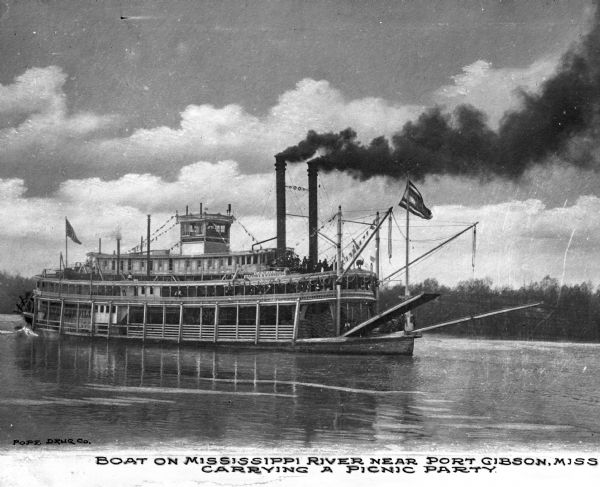 View of the steamboat "Senator Cordill" in the Mississippi River near Port Gibson. The boat is carrying an excursion party, and some of the passengers are on board. The name painted on the ship reads: "U.S. Mail Natchez & Vicksburg" and "Senator Cordill." Text on photograph reads: "Boat on Mississippi River near Port Gibson, Miss. Carrying a picnic party."