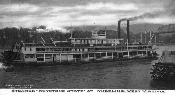 A view of the steamboat "Keystone State" on the Ohio River. "Keystone State" is painted on the rear of the ship. A small paddle ship is nearby, and the town and a bridge are in the background. Text on the photograph reads: "Steamer "Keystone State" at Wheeling, West Virginia."