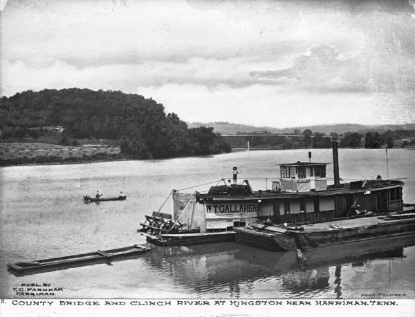 View of the steamboat "W.T. Gallaher" docked in the Clinch River. Two boys are on the boat, and a sign painted on the side and top reads: "W.T. Gallaher." "W.T. Gallaher Chattanooga" is on the rear of the ship. Two people are nearby in a canoe with a paddle. The County Bridge is in the background. Text on photograph reads: "County Bridge and Clinch River at Kingston near Harriman, Tenn."