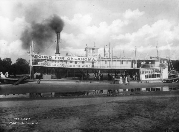 View of the docked steamboat "City Of Muskogee". "City Of Muskogee" is painted on the rear and top of the boat. Banners reading "Bound For Oklahoma" and "The Biggest Oil And Gas Field In The World" are displayed on the side of the ship. Several men and women pose on the boat and on shore.