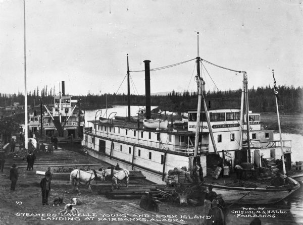 View of several steamboats docked. Boats in the foreground are the "Lavelle Young" and "Isabelle" (text "Isabelle" partially obscured). Boat in the background is the Rock Island. Cargo is either being loaded or unloaded from the "Lavelle Young." Many men are on shore, as well as a pair of horses and some dogs.
