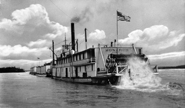 View of the paddle steamer "Monarch" and two barges in the river. "Monarch O.E.G." is painted on the rear of the ship. Two men are on the "Monarch" and on the top of one of the barges. An American flag is on the rear of the boat.