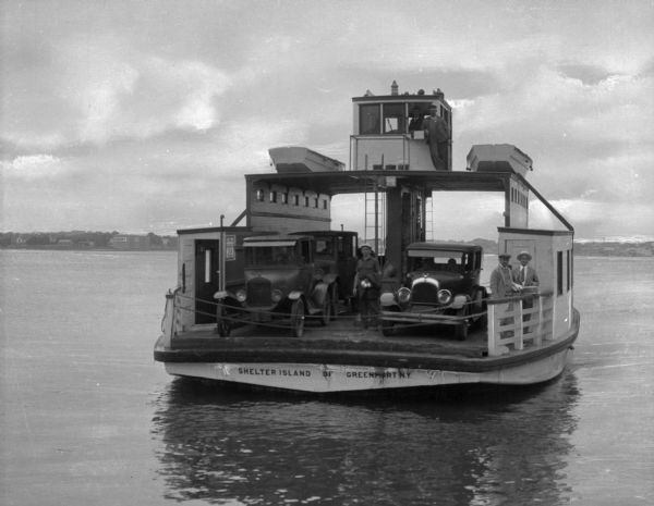 View of an automobile ferry boat traveling to or from Shelter Island Heights. Several men are on the ferry, along with three automobiles. "Shelter Island Of Greenport N.Y." is painted on the ferry.
