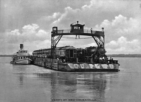 View of a barge transporting railroad cars and an engine across a river. Several men are on the barge, including one on an overhead structure. Text on photograph reads: "Ferry At New Orleans, LA."