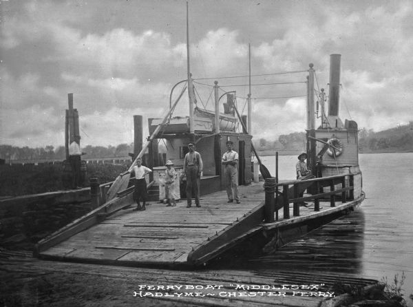 View of the ferryboat "Middlesex" docked in the Connecticut River. Two men and three boys stand on the boat, and another man are on shore. "Middlesex" is painted on a life preserver. Caption reads: "Ferryboat "Middlesex" Hadlyme-Chester Ferry."