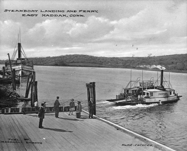 Elevated view of a ferry boat near a landing. "Gen. Spencer" is painted on the side of the ferry, which is transporting a cart, two children and a man. Several more children and men wait on the dock. A steamboat is docked nearby. Caption reads: "Steamboat landing and ferry, East Haddam, Conn."