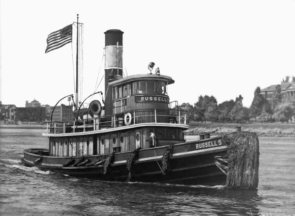 View of the "Russell 5" tugboat in the water near New York City. An American flag is flying on the boat. "Russell 5" is painted on the vessel. The shoreline and some buildings are in the background.