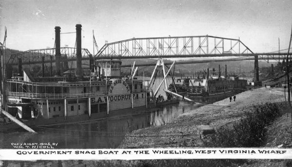 A view of the U.S. snagboat "E.A. Woodruff" at a wharf. "U.S. Snag Boat E.A. Woodruff" signs are on the side of the boat. A bridge and two paddle ships are nearby. Caption reads: "Government Snag Boat At The Wheeling, West Virginia Wharf."