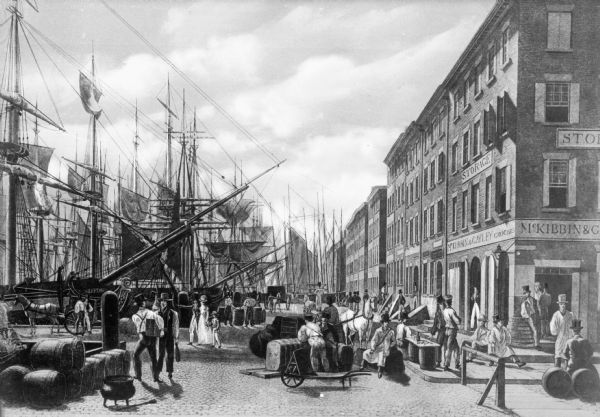 An illustration depicting an early scene at the wharf in New York City. Many sailing ships are along the left side of the illustration, and "Leeds" is the name of one ship. Pedestrians, cargo and horse-drawn vehicles are present on the busy street. Signs for "Storage" and "McKibbin & Gayley Grocers" are on a building on the right side of the street.