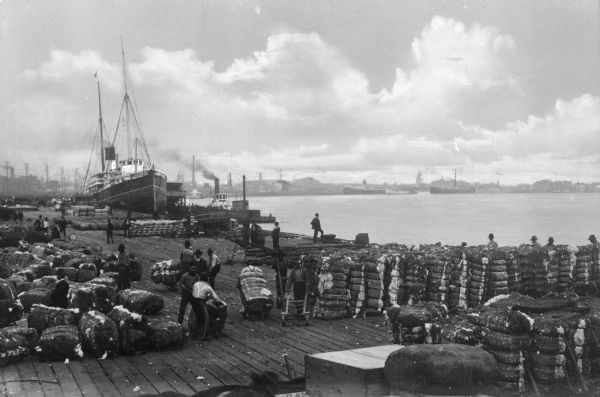 Elevated view of a dock scene, where workers are preparing bales of cotton for loading. A steamship and a tugboat are nearby. More ships and the shoreline are in the distance.