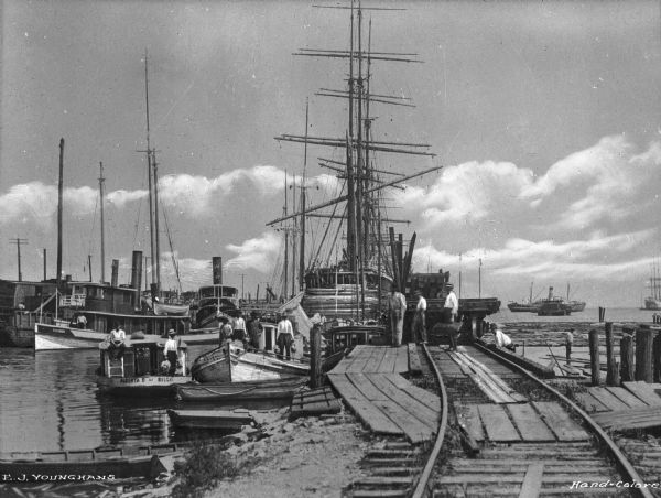 View of a shipyard with tracks for timber wagons. Several men are both on shore, and some men are on boats. One of the boats near the docks reads: "Alberta B. Of Biloxi" A boat in the background reads: "Eleanor" and another boat's smokestack reads: "W".