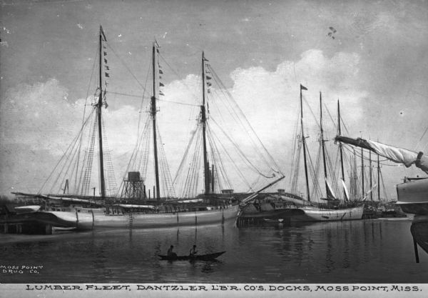 View of the fleet owned by Dantzler Lumber Company. Several large sailing vessels are docked, and nearby is a canoe with two passengers. Caption reads: "Lumber Fleet, Dantzler L'B'R. Co's Docks, Moss Point, Miss."