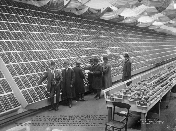 Two men pose while several others judge apples from Kelowna, British Columbia, Canada, at the national apple show in Vancouver, British Columbia, Canada. A large display wall filled with boxes of apples fills the exhibition hall. Nearby is a long table with apples. Banners are hanging overhead. Caption reads: "The Finest Car-Load Of Jonathans Ever Exhibited. Kelowna's Car-Load In The National Apple Show, Vancouver, B.C. The Judges In The Foreground Awarding First Prize, The Grand Sweepstakes."