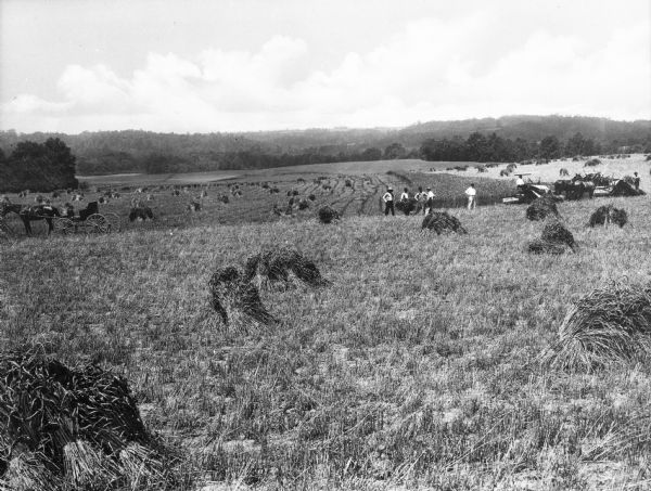 Two teams of horses aren pulling agricultural machinery, and a horse-drawn carriage is nearby. Most of the large field has been harvested, and bundles of wheat are scattered about.