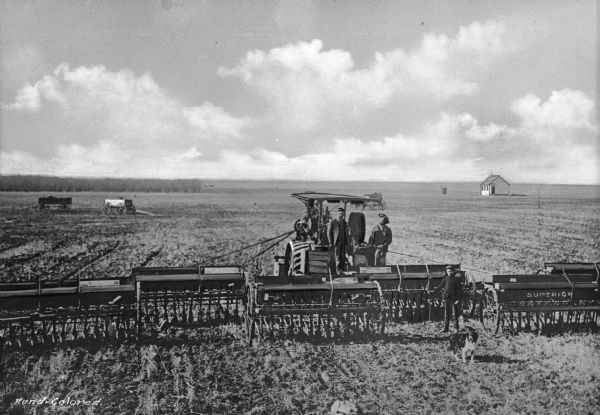Elevated view of two men using agricultural machinery to drill (plant) wheat. "Superior" sign is on the machinery. A boy and a dog stand nearby. A horse and cart and two other carts are  in the field. A small house is in the distance.