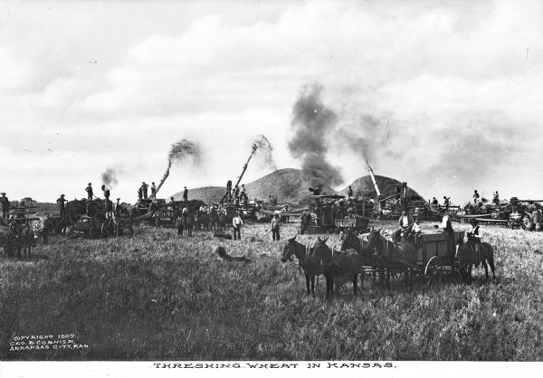 A large group of agricultural workers is shown threshing wheat in a field. In the foreground a horse-drawn vehicle is in the field. In the background are several horse-drawn wagons sitting near threshing machinery, which is piling processed wheat. Caption reads: "Threshing Wheat In Kansas."