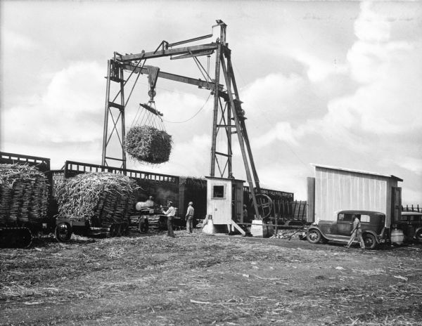 Several men loading sugar cane bales into train cars using a hoist. The text "USS Corp. 24" (partially obscured) is painted on one of the train cars.