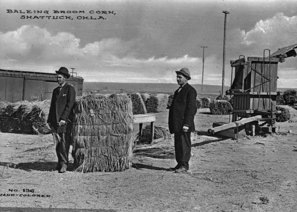 Two men standing near bound bales of corn stalks on a wharf. A train car is visible in the background. Caption reads: "Baleing[sic] Broom Corn, Shattuck, Okla."