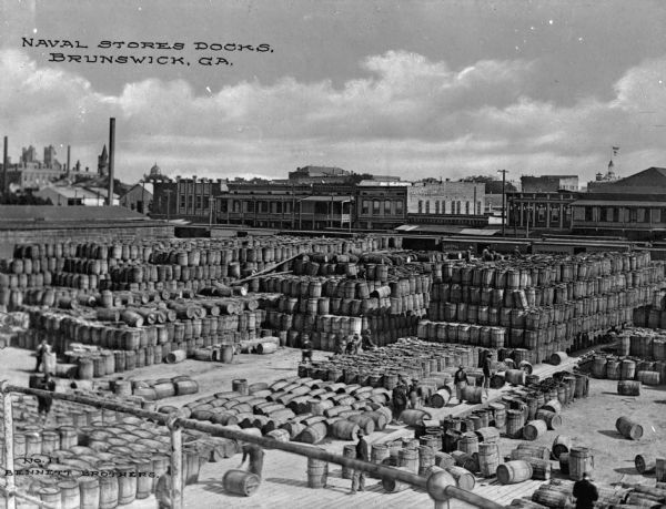 The naval stores docks piled high with barrels. Men can be seen rolling the barrels and buildings are in the background. Caption reads: "Naval Storage Docks, Brunswick, GA."