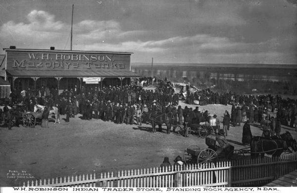 Elevated view of large crowd congregating with horse-drawn buggies outside of W.H. Robinson, an Indian trading store. Caption reads: "W.H. Robinson Indian Trading Store, Standing Rock Agency, N. Dak."