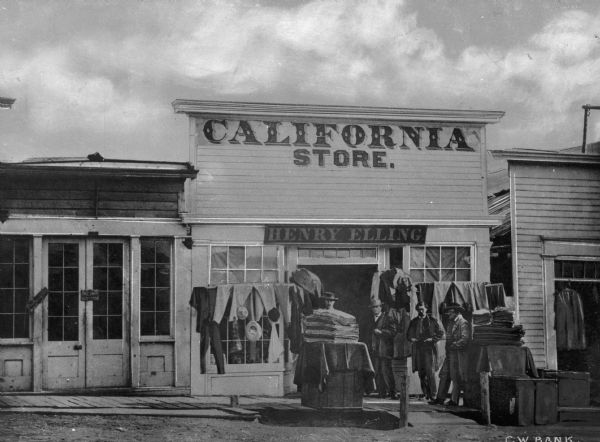 Men standing in front of the Henry Elling California general store, one of the first in Montana, with goods presented around the entry way.