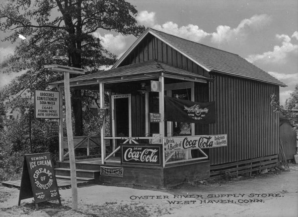 Exterior view of Oyster River Supply Store, a small rural general store selling groceries and other items. Signage reads "Groceries, Confectionery, Soda Water, Cigars, Fishing Tackle, Auto Supplies" "Semon's Ice Cream, Cream of Perfection" "Drink a Bottle of Coca-Cola, Delicious and Refreshing" "Polarine Motor Car Oil Sold Here." Caption reads: "Oyster River Supply Store, West Haven, Conn."