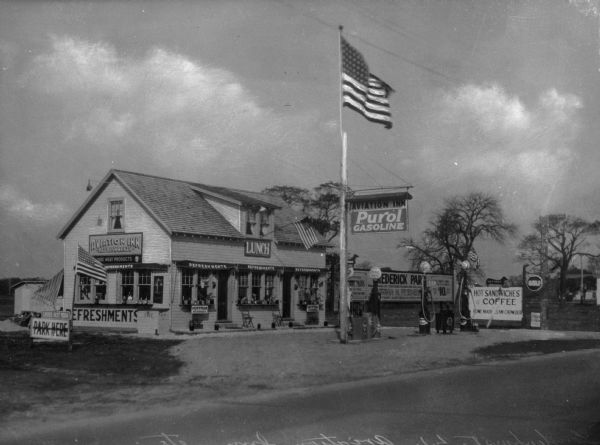 View across street toward the Aviation Inn filling station featuring two prominent American flags. Signage reads "Aviation Inn Refreshments" "Park Here" "Hoffman Beverages" "Purol Gasoline" "Hot Dogs. Hot Sandwiches and Coffee. Homemade Clam Chowder"