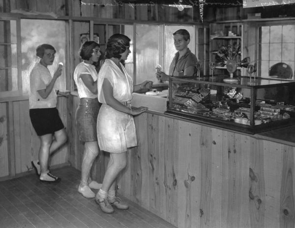 Camp-girls buying ice cream cones from a woman at the Camp Sherwood Shoppe. Candy bars and lollipops are on the counter, and a Camp Sherwood pennant is in the background.
