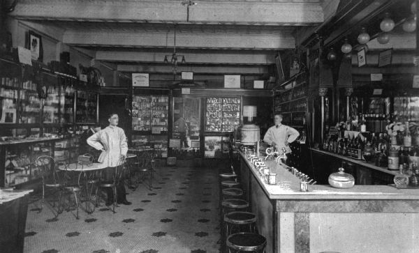 Interior of a drugstore with a soda fountain, seating, and two men staffing the counter. Items and signage lines the walls.