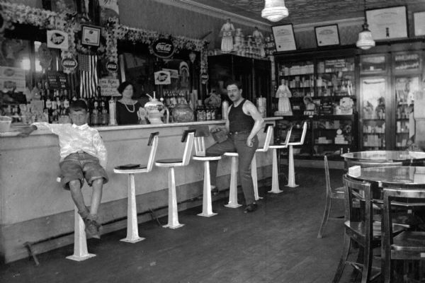 Interior view of the Midland Beach Park Drug Co. soda shop. A woman stands behind the counter near a soda fountain, while a man and a young boy lounge at the bar. Candy is for sale and there are a number of signs on the wall.