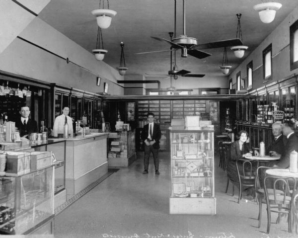 Interior view of a drug store with staff standing behind the counter and customers seated at tables. Goods are on display and the store features overhead fans.