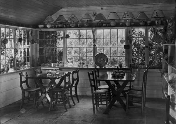 Most likely the interior of a gift shop featuring two sets of tables and chairs arranged in front of a large window with a number of similar lamps lining a shelf above the window.
