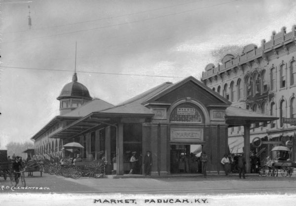 A covered outdoor market of Grecian design in Paducha, KY. Men stand around outside and a horse-drawn cart is visible to the right of the Market. Sign above entrance reads "Wagons Not Allowed to Stand at This End of the Market House." Caption reads: "Market, Paducah, KY."