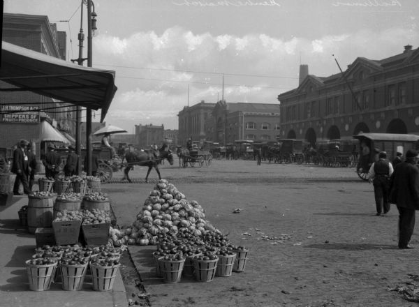 An open air fruit stand in the Center Market in Baltimore, MD. What appears to be cabbages, apples, potatoes and onions are in piles or in baskets. Horse-drawn carriages and pedestrians are visible in the background navigating the broad brick-laid roads. Signage reads "J.H. Thompson & Co. [illegible] Shippers of Fruit and Produce."