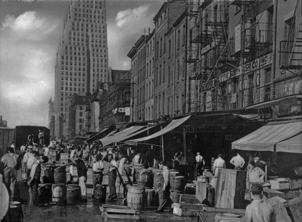 An open air market in the streets of New York. Barrels of goods line the sidewalk and behind them fire-escapes can be seen going up the sides of brick tenement buildings. Far in the background a tall building is visible.