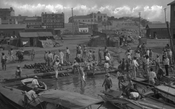 Elevated view of a number of people unloading goods from boats arriving at a waterfront marketplace. Huts and various other buildings are visible on the hillsides in the background.