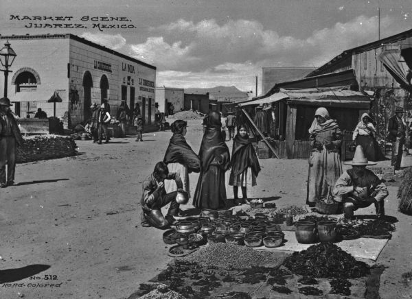 A market scene in Juarez, Mexico with people standing around pottery and various other items being sold from a blanket laid on the ground.