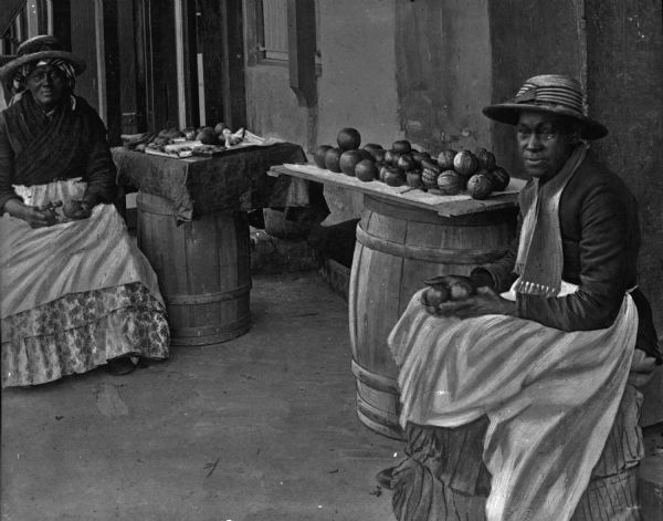Two African American women wearing hats selling fruit at a stand made of wood panels placed on top of barrels in Bermuda.