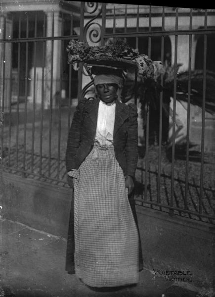 African American vegetable vendor with a large basket of vegetables balanced on her head.