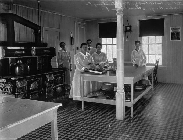 Interior view of kitchen at the Hotel Hopp "Where Good Things Come From." Kitchen staff of four women and one man are posed behind a large table. In the background to the left is a large cast iron oven.