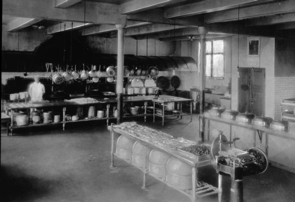 Interior view of kitchen at the Grand Lodge Hall with a male cook posed behind a large table underneath a hanging rack of pots and pans. There's a large stove behind the cook.