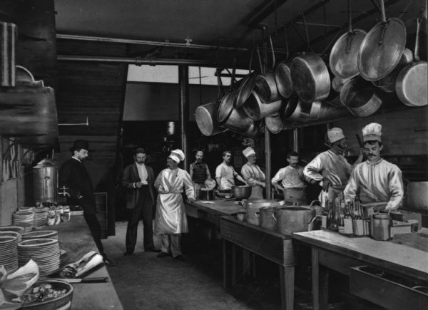 Kitchen workers at the Hotel Astor. Men standing around long tables with a hanging rack of pots and pans over a long table. Plates are piled up on another table to the left of the image.
