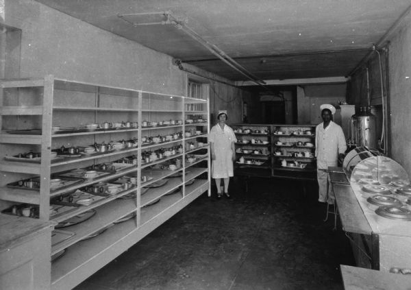 Two kitchen workers at Mary's Sanitorium stand in a kitchen room with shelves of trays and a warming tray along the walls.