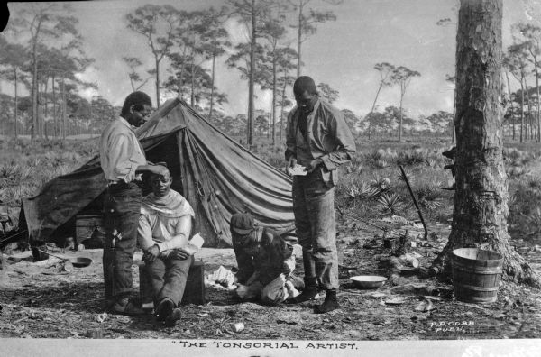 Four African-Americans with a tent in the background. One man is getting his hair cut. Caption reads: "The Tonsorial Artist."
