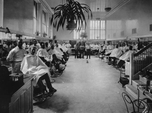 Interior view of the Union Station Barber Shop with a number of men seated in chairs with barbers standing behind them. Plants and light fixtures are suspended from the high ceiling.