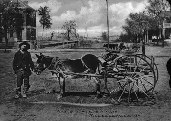 A man standing in the middle of a dirt road next to a small mule-drawn cart laden with chopped wood. Caption reads: "'Any Splinters Today' Milledgeville, GA."