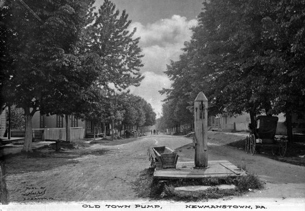 View of a water pump in the middle of a dirt road. In the background a horse-drawn buggy is parked on the side of the road. A small group of people are in the background. Caption reads: "Old Town Pump, Newmanstown, PA."