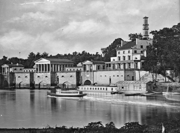 Exterior of Fairmount Water Works with numerous columned buildings overlooking a river with boats moored to a small dock. Another boat is on the river in motion.