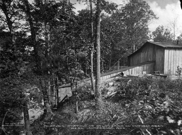 Exterior of a wooden water pumping station in a wooded area. Caption reads: "Pumping Station at Stuttgart on Cotton Belt, Near Jonesboro, Ark."